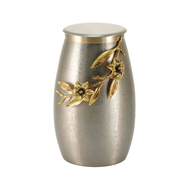 A brushed silver metal urn with a gold-tone floral branch and flower decoration on the front, set against a plain white background.